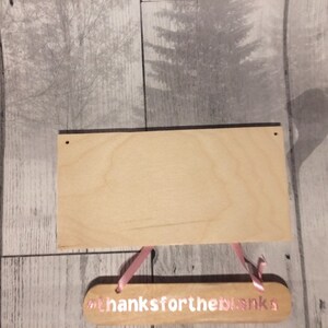 May include: A blank wooden sign with two holes at the top for hanging. The sign is attached to a smaller wooden sign with pink ribbon and the text "#thanksfortheblanks".