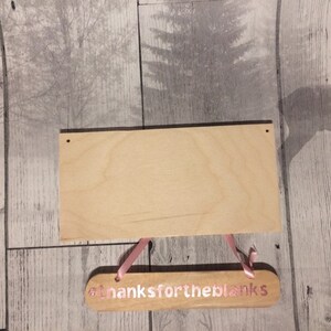 May include: A blank wooden sign with two holes at the top and a smaller wooden sign hanging below it. The smaller sign has the text "thanks for the blanks" in pink lettering.