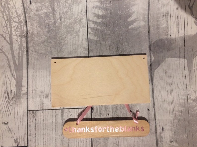May include: A blank wooden sign with two holes at the top and a smaller wooden sign hanging below it. The smaller sign has the text "thanks for the blanks" in pink lettering.