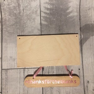 May include: A wooden sign with the text "Thanks for the blanks" in pink lettering. The sign is hanging from a pink ribbon and is attached to a larger, blank wooden sign.