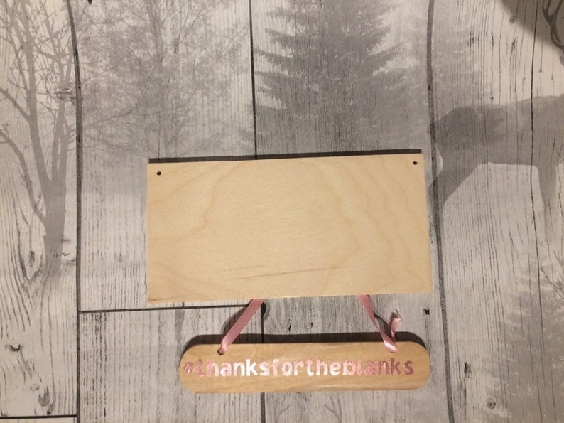 May include: A wooden sign with the text "Thanks for the blanks" in pink lettering. The sign is hanging from a pink ribbon and is attached to a larger, blank wooden sign.