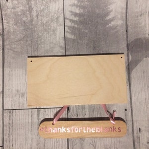 May include: A wooden sign with the text "Thanks for the blanks" in pink lettering. The sign is hanging from pink ribbon and is on a woodgrain background.