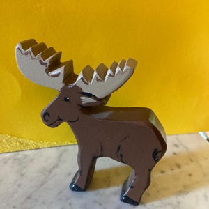 May include: A brown wooden moose figurine with large, light tan antlers. The moose has a painted black eye, a simple black line for a mouth, and black painted hooves. The body is a solid brown color with visible brush strokes and a smooth finish.