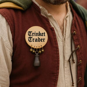 May include: A wooden Trinket Trader badge with a black inscription and a small bell hanging from it. The badge is attached to a burgundy vest over a cream-colored shirt. The vest has a green hood.