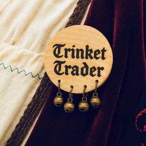 May include: A circular wooden sign, inscribed with "Trinket Trader" in black lettering, features four small brass bells suspended below. The background includes cream fabric, a dark brown braid, and a deep red velvet material.
