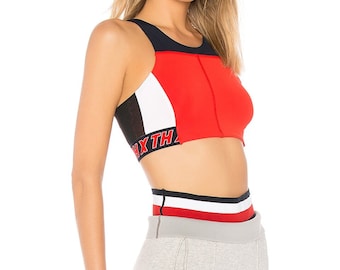 Puede incluir: Un sujetador deportivo rojo, blanco y negro con el texto "NX TH" en la banda. El sujetador se combina con pantalones de chándal grises. El conjunto está diseñado para actividades deportivas.