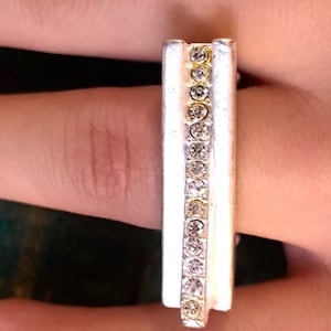 May include: A silver-toned ring with a rectangular design. The ring features a central row of clear, sparkling rhinestones, flanked by two silver bands. The ring is worn on a finger, showcasing its elegant and simple design.