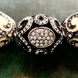 May include: Three decorative egg-shaped beads. The beads have a cream-colored base with black scrollwork and are encrusted with sparkling rhinestones. The center bead is black with a rhinestone center.