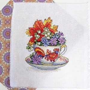 May include: A white teacup with a rooster design and a bouquet of fall flowers, including red, orange, yellow, white, and purple flowers. The teacup is on a white saucer with a floral pattern.