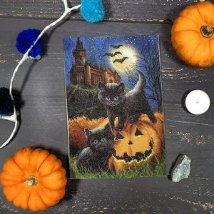May include: A cross-stitch design featuring two black cats perched on a jack-o'-lantern with a spooky castle in the background. The design is set against a dark blue night sky with a full moon and bats.