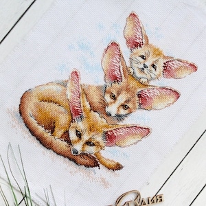 May include: A cross-stitch embroidery of three fennec foxes with large ears. The foxes are depicted in a light brown color with pink ears and are arranged in a stacked formation. The background is a light blue color.