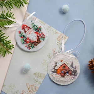 May include: Two handmade Christmas ornaments, one with a red and white gnome and the other with a red house in a snowy forest. Both ornaments are round and white with beaded details.