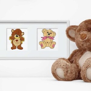 May include: Three framed cross-stitch patterns of teddy bears. The first bear is brown and wearing a yellow shirt. The second bear is brown and holding a heart-shaped sign. The third bear is light brown and wearing a pink bow. A large brown teddy bear with a white bow is sitting in front of the frames.