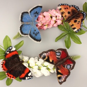 May include: Four felt butterflies in different colors and patterns, including blue, brown, red, and orange, are displayed on a gray background with white and pink flowers.