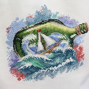 May include: A white pillow with a green glass bottle with a cork stopper. The bottle contains a small white sailboat on a blue and green ocean. The bottle is surrounded by a watercolor-like design of blue, green, red, and white.