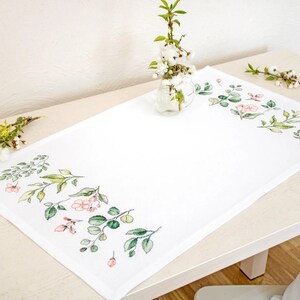 May include: White table runner with a delicate floral design featuring pink flowers and green leaves. The runner is on a wooden table with a vase of white flowers in the background.