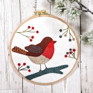 May include: Embroidered hoop art featuring a red-breasted robin perched on a branch with green leaves and red berries. The background is white felt.