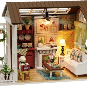 Puede incluir: Una casa de mu&ntilde;ecas en miniatura con un exterior blanco, un techo marr&oacute;n y una valla de madera blanca. El interior est&aacute; decorado con una chimenea, un sof&aacute;, una mesa de caf&eacute; y una peque&ntilde;a figura de perro. La casa de mu&ntilde;ecas tiene un techo de cristal y una l&aacute;mpara.