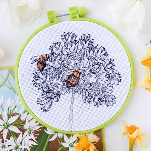 May include: A cross-stitch embroidery hoop featuring a floral design with two bees. The embroidery is in black and white with orange and brown accents. The hoop is green, and the background includes white tulips and yellow daffodils.