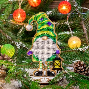 May include: A green and purple gnome with a white beard and a lantern, standing on a wooden base. The gnome is decorated with beads and is a Christmas ornament.