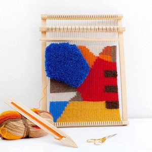 Puede incluir: Un telar de madera con un tapiz tejido de colores con formas geom&eacute;tricas en tonos azules, rojos, amarillos y marrones. Tambi&eacute;n se incluye una bola de hilo naranja y beige y una aguja de madera.