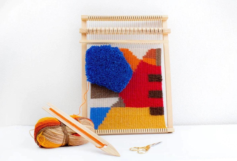 Puede incluir: Un telar de madera con un tapiz tejido de colores con formas geom&eacute;tricas en tonos azules, rojos, amarillos y marrones. Tambi&eacute;n se incluye una bola de hilo naranja y beige y una aguja de madera.