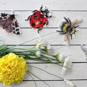 May include: Five embroidered insect designs on a white wooden background. The designs include a moth, ladybug, bumblebee, dragonfly, and a yellow flower.