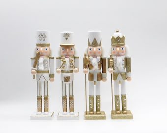 May include: A set of four decorative nutcracker figurines standing in a row. Each figure is painted in a white and gold color scheme with glitter accents. They feature various headgear including hats and crowns, and hold different accessories like a sword, drumsticks, and staffs. The figures are uniform in style, showcasing detailed facial features and ornate uniforms.