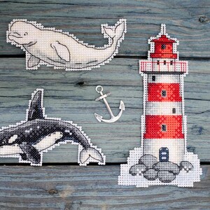 May include: Four cross-stitch embroidery designs on a wooden background. The designs include a white beluga whale, a black and white orca whale, a red and white lighthouse, and a silver anchor.