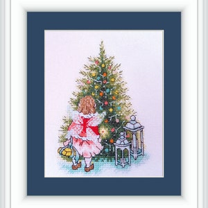 May include: Cross-stitch artwork of a young person in a red dress with a white bow, standing before a decorated Christmas tree. The artwork includes two white lanterns and is framed in white with a navy blue mat.