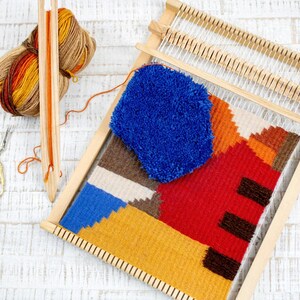 Puede incluir: Un telar de madera con un tapiz tejido de colores con formas geom&eacute;tricas en tonos rojos, naranjas, amarillos, azules y marrones. Tambi&eacute;n se puede ver una madeja de lana azul y un par de tijeras.