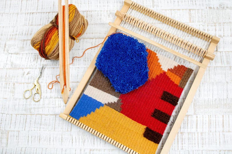 Puede incluir: Un telar de madera con un tapiz tejido de colores con formas geom&eacute;tricas en tonos rojos, naranjas, amarillos, azules y marrones. Tambi&eacute;n se puede ver una madeja de lana azul y un par de tijeras.