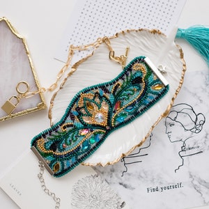 May include: A beaded bracelet with a blue, green, and gold design. The bracelet is on a white seashell-shaped dish with a gold trim. The bracelet has a silver clasp and a chain.