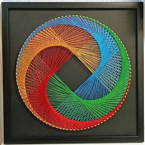 May include: A square black frame containing circular string art. The design features vibrant red, orange, yellow, blue, and green threads wrapped around gold-coloured pins, creating a geometric, swirling pattern against a solid black background.