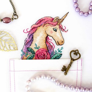 May include: A white fabric pocket with pink stitching, a pink and purple unicorn cross-stitch design, a gold key, and two pearl necklaces.