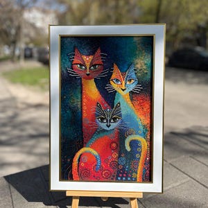 May include: A framed diamond painting featuring three stylized cats in vibrant shades of red, orange, blue, and yellow. The artwork displays a dark, speckled background with swirling patterns and is encased in a white frame with a thin gold inner border.