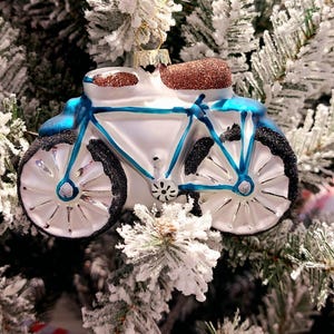 May include: A bicycle-shaped Christmas ornament. The ornament is white with blue accents and black wheels. The seat and handlebars are covered in brown glitter. The ornament is hanging on a snow-covered Christmas tree.