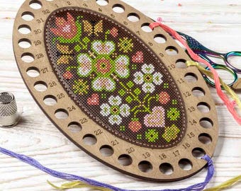 May include: Wooden embroidery floss organizer with a floral cross-stitch design. The oval-shaped organizer has numbered holes around the edge for thread. Colorful embroidery floss and scissors are also visible.