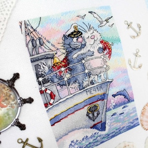 May include: A cross-stitch embroidery of two cats on a boat. The cat in the captain's hat is holding a white cat with a red scarf. The boat is blue and white with the word "MEЧTA" written on the side. The background is a colorful sunset.