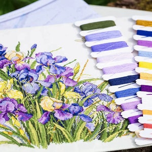 Puede incluir: Un patr&oacute;n de punto de cruz que presenta un ramo de iris p&uacute;rpuras, azules y amarillas. El patr&oacute;n va acompa&ntilde;ado de un juego de hilos de bordar en varios colores, incluyendo morado, azul, amarillo, naranja y verde.