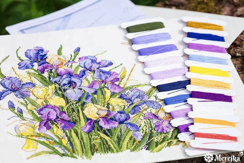Puede incluir: Un patr&oacute;n de punto de cruz que presenta un ramo de iris p&uacute;rpuras, azules y amarillas. El patr&oacute;n va acompa&ntilde;ado de un juego de hilos de bordar en varios colores, incluyendo morado, azul, amarillo, naranja y verde.