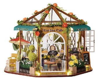 Puede incluir: Un modelo de casa de muñecas de madera en miniatura de una cafetería de jardín con un toldo verde, un techo de vidrio y un letrero que dice "Garden Cafe". La cafetería tiene una pequeña terraza con una mesa y sillas, y una variedad de plantas y flores.