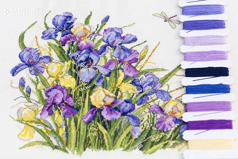 Puede incluir: Patr&oacute;n de punto de cruz con un ramo de iris p&uacute;rpuras, azules y amarillas con hojas verdes y una lib&eacute;lula. El patr&oacute;n va acompa&ntilde;ado de un juego de hilos de bordar en varios tonos de p&uacute;rpura, azul y amarillo.