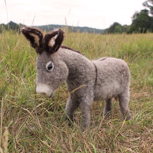 May include: A handcrafted felt donkey figurine in a grassy field. The donkey is primarily gray with darker brown accents on its ears and mane. It has a white muzzle and is standing in tall grass, with a blurred background of trees and sky.