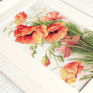 May include: Cross-stitch artwork of a bouquet of vibrant red poppies with green foliage and white accents, framed in a white wooden frame. The detailed needlework showcases the flowers' textures and colors.