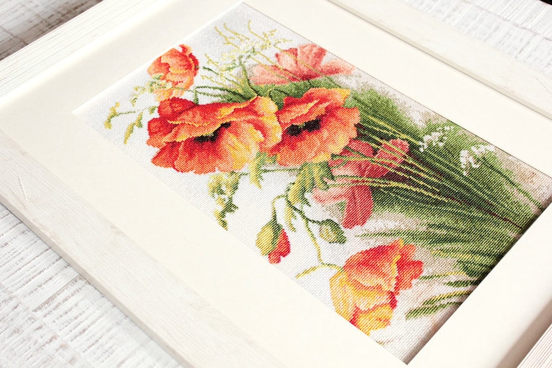 May include: Cross-stitch artwork of a bouquet of vibrant red poppies with green foliage and white accents, framed in a white wooden frame. The detailed needlework showcases the flowers' textures and colors.