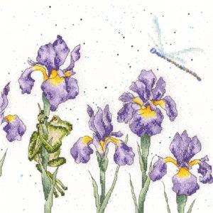 May include: Cross-stitch design featuring purple irises with yellow centers, a green frog, and a dragonfly against a white background. The needlework showcases detailed floral and animal elements, with a delicate, handcrafted aesthetic.