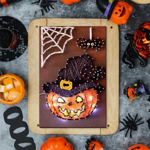 May include: A Halloween themed string art piece with a pumpkin wearing a witch's hat. The pumpkin is lit up with white lights and has a scary face. The string art is surrounded by other Halloween decorations, including pumpkins, spiders, and candy.