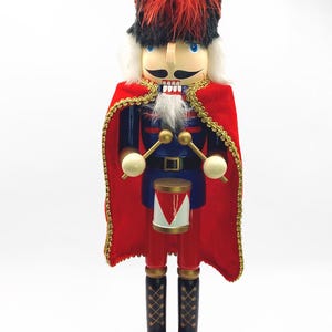 May include: A wooden nutcracker soldier in a red and blue uniform with a red cape and a tall black hat with red feathers. The nutcracker has a white beard and mustache, and holds drumsticks. The base is red.