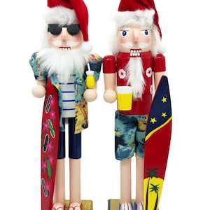 May include: Two nutcracker figures wearing red Santa hats and beach attire. The left figure wears sunglasses, a tropical shirt, and holds a surfboard and drink. The right figure wears a red shirt, patterned shorts, and holds a surfboard and cup.
